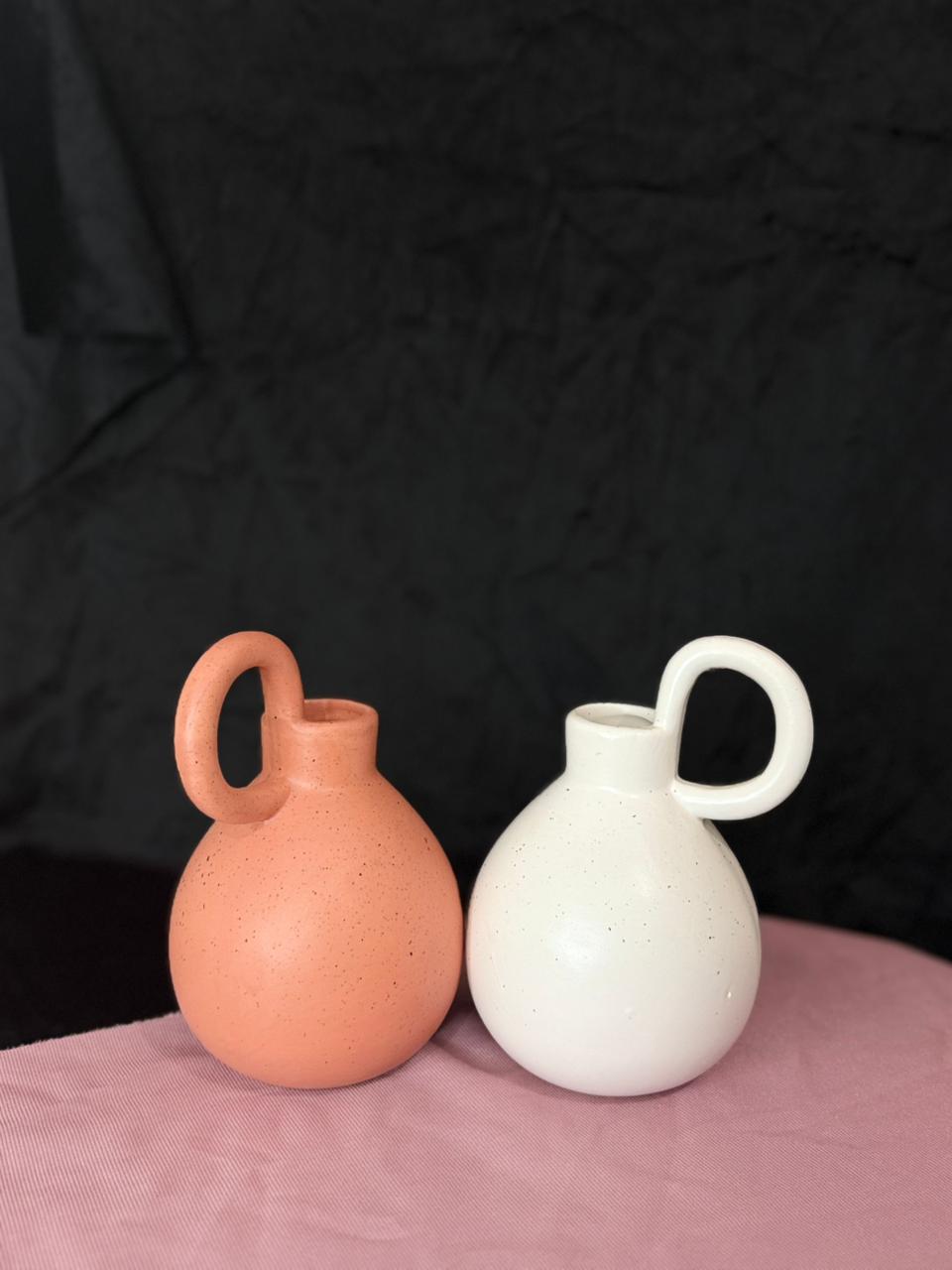 Two ceramic vases, one terracotta and one white, on a pink surface with a black background.
